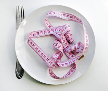 Beyaz arka planda beyaz plaka, çatal ve pembe mezura. Kilo verme konsepti, bembeyaz bebek. Arka planın üst görüntüsü. Beyaz arkaplandaki uzunluk ve genişliği ölçmek için santimetre. Tasarımınız için terzi mezurası.