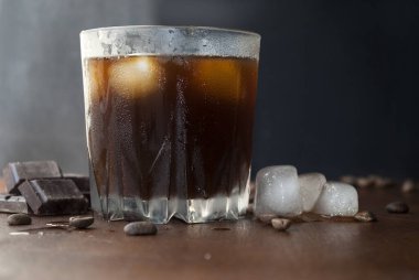 Buzlu kahve, bir bardak soğuk kahve. Koyu çikolata ve kahve çekirdeği. Yan görünüm, koyu arkaplan.