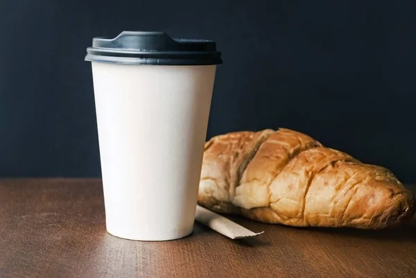 Coffee to go in a paper cup with croissants on a wooden table.