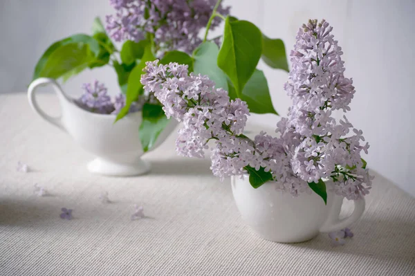 Room interior with gentle purple lilacs flower blossom in jug and tea cup on table with linen tablecloth, tender romantic spring home decor in morning light, decorating house with syringa plant.