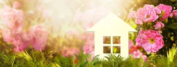 Eco friendly house model in green grass and rose flowers in garden, ecological sustainable lifestyle, life harmony. Buying or selling real estate, investment concept, wide panoramic banner.