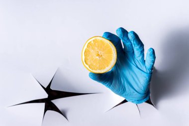 Kağıt duvardaki bir delikten bir kadın eli, mavi tıbbi eldivenlerle parlak sarı bir limon tutuyor.