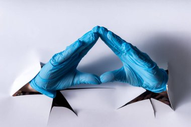 Beyaz kağıttan yapılmış yırtık bir duvarın içinden geçen mavi eldivenli bir kadın. El işareti, kendini izole etme. Duvardaki delik. jest enfeksiyonu salgını