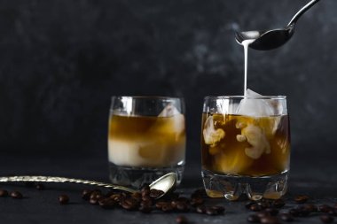 Bir bardak soğuk ve lezzetli aromalı kahveye koyu arkaplanda buzla süt dökmek.