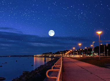 Limandaki yıldızlı gökyüzünde ay, gece bulanık şehir ışığı, denizde pembe gün batımı, ufuktaki gemi insanlar gezinti bulvarında yürüyor, yazın bulutlu akşam gökyüzü, doğa manzarası.