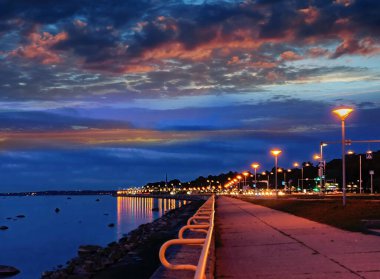 Şehir gece limanda bulanık ışık, denizde pembe mavi altın rengi günbatımı, deniz kenarındaki insanlar gezinti bulvarında yürüyor, yazın bulutlu akşam gökyüzü, doğanın romantik arka planı 