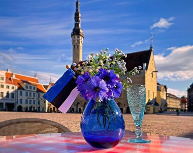 Tallinn eski kasaba meydanında Estonya ulusal bayrağı, mavi vazoda mavi bir çiçek buketi sembolleri ve başkent kafeteryada bir bardak şampanya, Estonya seyahatini ziyaret edin