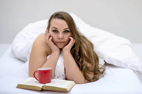 Çekici, halinden memnun, genç, kilolu, kilolu, tombul, seksi, uzun saçlı bir kadının portresi. Yatakta rahatça uzanan kitap ve kahvesini taze, yumuşak, beyaz bir bardakta içiyor..