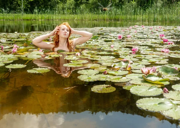 美しい若いセクシーな赤髪の女性人魚は 官能的かつ魅惑的に水 ピンクの睡蓮と伸び 彼女の髪をストローク — ストック写真