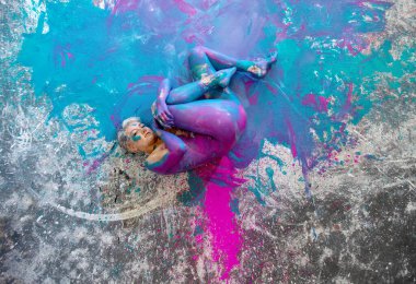 Young naked woman in turquoise and magenta paint color, painted, lies decorative, elegantly curled up in fetus pose on the colorful studio floor. Creative, abstract expressive body art and painting. clipart