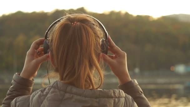 Das entspannte rothaarige Mädchen setzt Kopfhörer auf und genießt die Musik auf dem Hintergrund des Flusses im Freien bei Sonnenuntergang. lauscht den Geräuschen der Natur. Schallheilungskonzept. Entspannung — Stockvideo