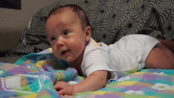 Adorable niño feliz de tres meses de edad acostado boca abajo en una manta a cuadros en la cama y sonríe. Muestra la lengua con la boca abierta. Calcetín en una pierna. Patas enrollables — Vídeo de stock