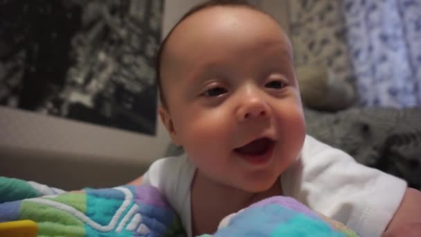 Funny cute little caucasian baby boy with brown eyes and dark hair lies on the parents bed and smiles at the camera and blowing bubbles and drool. Soft focus background. — Stock Video