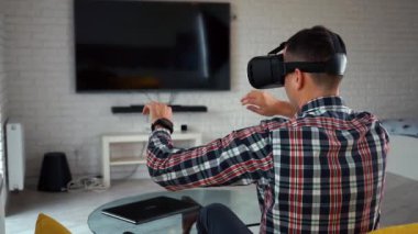 Vr-kulaklıklı genç, modern beyaz adam. Arkadan bak. Pencerenin yanındaki rahat, akıllı bir dairede, kanepede oturuyor. Artırılmış gerçeklikte bir bilgisayar oyunu oynuyor. Akıllı Ev. Ekose gömlek. Kamera açıklığı