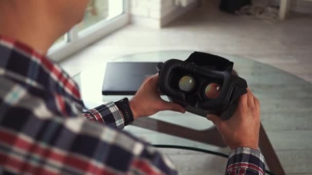 Young modern caucasian man puts on VR-headset. View from the back. Sitting on the couch in a cozy smart apartment near the window. Close-up of VR glasses helmet. Augmented reality — ストック動画