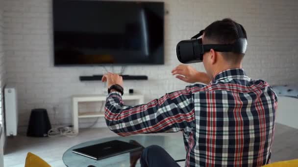 Joven hombre caucásico moderno con auriculares VR. Vista trasera. Se sienta en un sofá en un acogedor apartamento inteligente cerca de la ventana. Juega un juego de ordenador en realidad aumentada. Casa Inteligente. Camisa a cuadros. Espacio de la cámara — Vídeo de stock