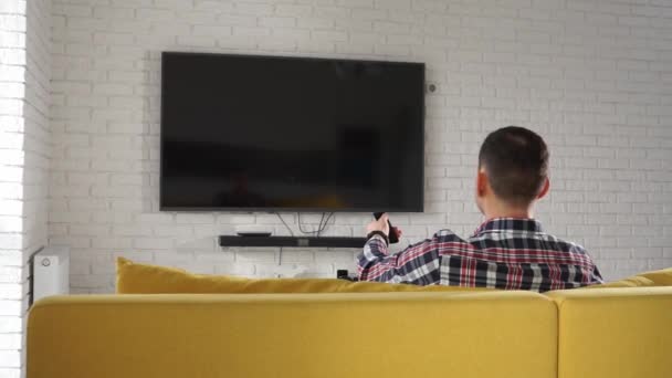 Junger moderner kaukasischer Mann im karierten Hemd, der auf dem gelben Sofa vor dem Plasma-Fernseher sitzt. Bildschirm ist schwarz. schaltet Kanäle mit der Fernbedienung. zurück — Stockvideo