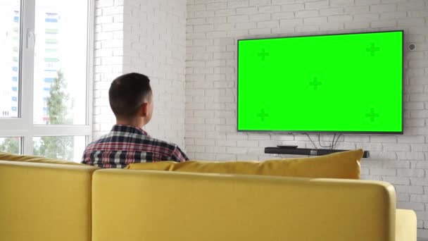 Joven hombre caucásico moderno en camisa a cuadros sentado en el sofá amarillo viendo televisión de plasma con cromakey pantalla verde solo en casa. Conmuta los canales usando el control remoto. Acogedora casa inteligente — Vídeo de stock