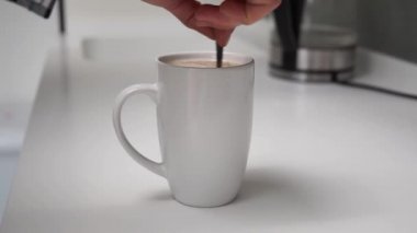 Beyaz kahve fincanındaki şekeri, beyaz kaşıklı sütlü sütlü sütlü sütlü, modern beyaz, yüksek teknolojili bir mutfakta veya kafede el pençe divan duran kişinin yan görüntüsü. Kapuçino ya da fincanda çay.
