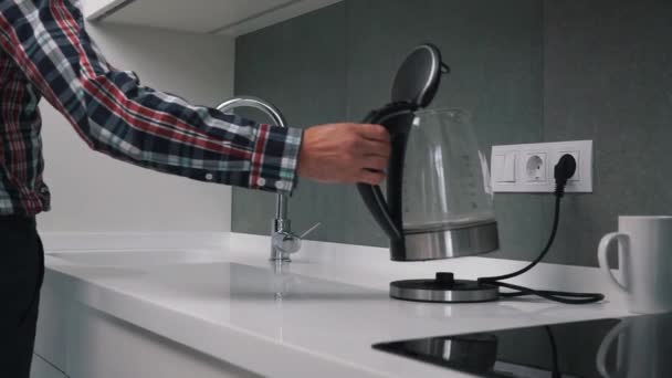 Man Giet water in glas transparante elektrische warm water thee ketel pot in een moderne witte high-tech keuken in een slimme home loft appartement. Kookt water om thee of koffie te zetten — Stockvideo