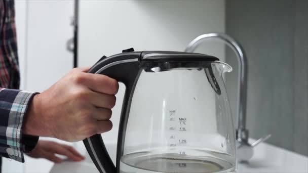 L'uomo accende il bicchiere trasparente elettrico bollitore per tè acqua calda in una moderna cucina bianca high-tech in un appartamento loft casa intelligente. Bollisce l'acqua per fare il tè o il caffè — Video Stock