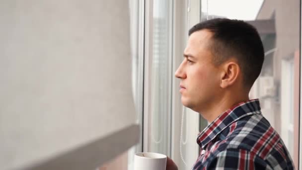 Joven hombre de negocios guapo camina hacia la ventana con persianas o jalousie. Empresario autónomo bebe café o té y mira por la ventana. En cámara lenta. Vista lateral — Vídeos de Stock