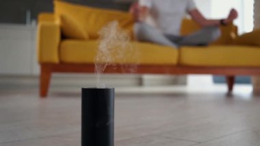 Genç, modern Kafkasyalı adam sarı bir kanepede oturmuş yoga nilüferleri yapıyor. Evde meditasyon yapmak, yatıştırma ve sakinleştirmenin tadını çıkarmak. Ahenkli sağlık yaşam tarzı. Hava nemlendirici ön planda.