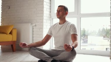 Genç, modern Kafkasyalı adam sarı bir kanepede oturmuş yoga nilüferleri yapıyor. Adam evde meditasyon yapıyor. Yatıştırma ve yatıştırmanın tadını çıkarıyor. Ahenkli sağlık yaşam tarzı. Gençlik ve meditasyon kavramı.