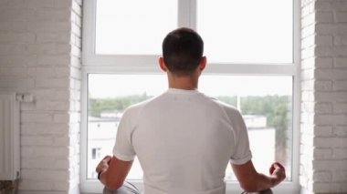 Genç modern Kafkasyalı adam pencerenin önünde oturmuş yoga nilüfer pozu çalışıyor. Adam evde meditasyon yapıyor. Yatıştırma ve yatıştırmanın tadını çıkarıyor. Ahenkli sağlık yaşam tarzı. Arka plan