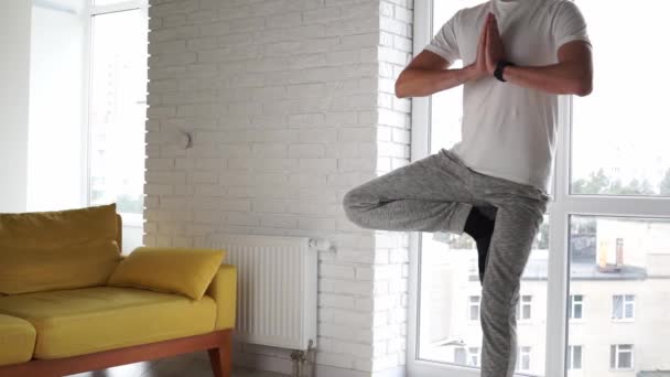 Jovem homem caucasiano moderno prática de ioga está em um pé equilibrando palmas juntos. Homem meditando em casa desfrutando de apaziguamento e pacificação. Harmonia estilo de vida bem-estar. Conceito de meditação . — Vídeo de Stock