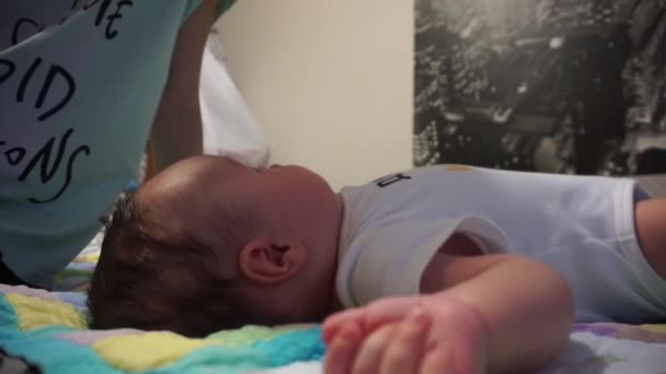 茶色の目と黒い髪をした小さな白人の赤ん坊は、入浴後に両親のベッドの上に彼の背中に横たわっています。ママは頭の上で赤ちゃんをパスします. — ストック動画