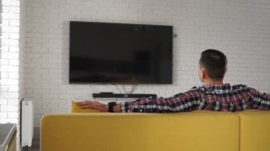 Young modern caucasian man in plaid shirt sitting on the yellow sofa watching plasma TV. Screen is black. Back view