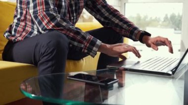 Close-up of hands typing on keyboard. Young man freelancer student sitting on the sofa in cozy room. Successful thoughtful millennial guy works at a laptop at a glass desk