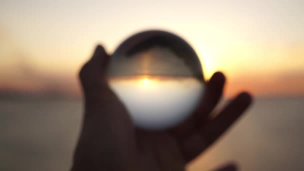 Το αρσενικό χέρι κρατά μια γυάλινη μπάλα σε φόντο ηλιοβασιλέματος στη θάλασσα. Δραματική κινηματογραφική λήψη. Σιλουέτα. — Αρχείο Βίντεο