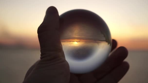 A férfi keze egy üveggolyót tart a naplemente hátterében a tengeren. Drámai filmfelvétel. Sziluett. — Stock videók