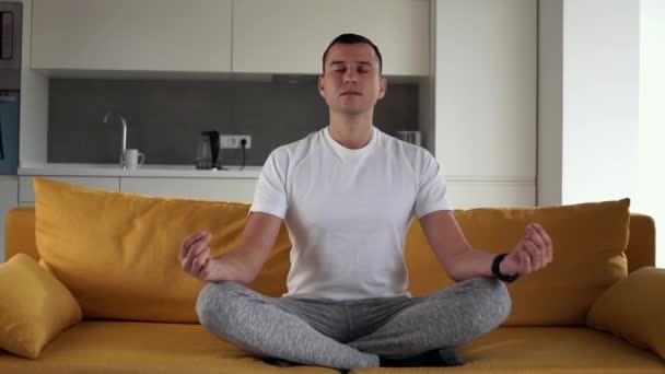 Junger moderner kaukasischer Mann praktiziert Yoga-Lotus-Pose sitzend auf einer gelben Couch. Mann, der zu Hause meditiert und Beschwichtigung und Befriedung genießt. Harmonie Wellness Lifestyle. Jugend und Meditationskonzept. — Stockvideo