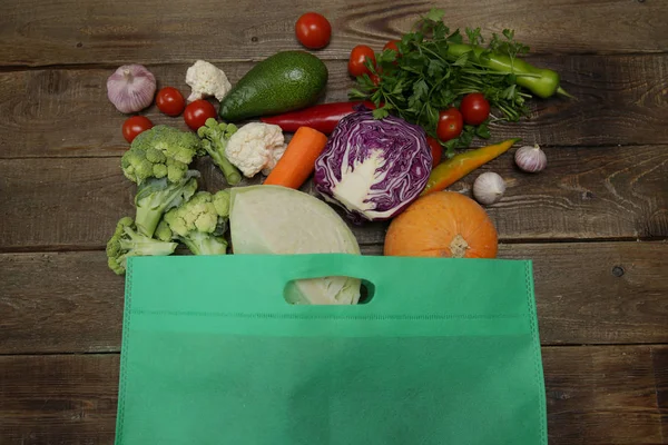 Organic fresh vegetables in a green bag on a wooden background: Broccoli, carrots, avocado, tomatoes, garlic, cauliflower, cabbage, parsley, pepper, pumpkin. Healthy food concept. Vegan. Copy space