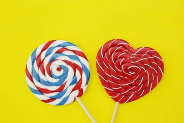 Kırmızı kalp şeklinde lolipoplar ve sarı arka planda izole edilmiş kırmızı-mavi-beyaz spiral. Şekerleme arka planı. Tatil ve hediye kavramı. Metin için kopyalama alanı ile.