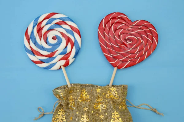 lollipops in the form of a red heart and a red-blue-white spiral and sugar candy cane in a gift burlap bag isolated on a blue background. Confectionery background. The concept of holidays and gifts.