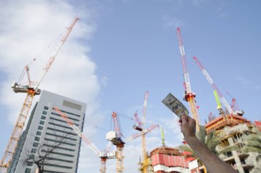 Vinçlerin ve bina inşaatının arka planında 100 Amerikan doları banknotları olan bir el. İnşaat endüstrisi. İş ve finans ve yatırım kavramı.