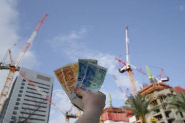 hand with one hundred and two hundred shekel banknotes from Israel on the background of a cranes and building construction. Construction industry. Concept of business and finance and investment. clipart
