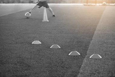 Siyah beyaz görüntü. Belirleyici koniler için seçici odak noktası, arka planda bulanık çocuk oyuncuların antrenman yaptığı yeşil yapay sahada futbol eğitim ekipmanları. Eğitim sınıfı için malzeme.