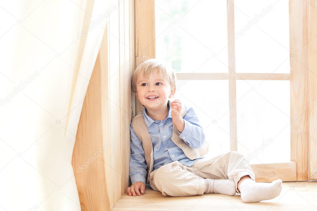 A little boy is sitting on the windowsill in the nursery. The concept of leisure, leisure, people and lifestyle. Portrait of a smiling boy sitting on a wooden window sill, looking at the camera.