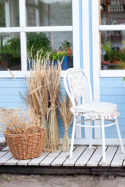 corner of garden in fall. Romantic porch, patio, chair and basket with spikelets near wooden blue house in countryside. Terrace in rustic style. Old coffee terrace, street cafe. Modern interior design