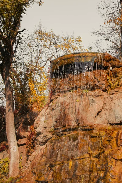 Waterfall from stones in the park. Against the background of autumn foliage and trees of orange and yellow. — Stock Photo, Image