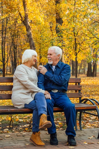 Feliz anciano y mujer sentados en un banco en el día de otoño. Pareja mayor relajada sentada en un banco del parque. Abuelo besa suavemente a la abuela en la frente . — Foto de Stock