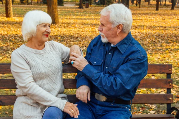 Happy elderly man and woman sitting on a bench in autumn day. Relaxed senior couple sitting on a park bench. Grandfather gently kisses grandmother on the forehead.