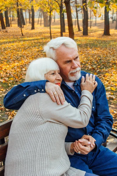 Glückliche ältere Männer und Frauen, die an einem Herbsttag auf einer Bank sitzen. Entspanntes Seniorenpaar auf einer Parkbank sitzend. Großvater küsst Großmutter sanft auf die Stirn. — Stockfoto