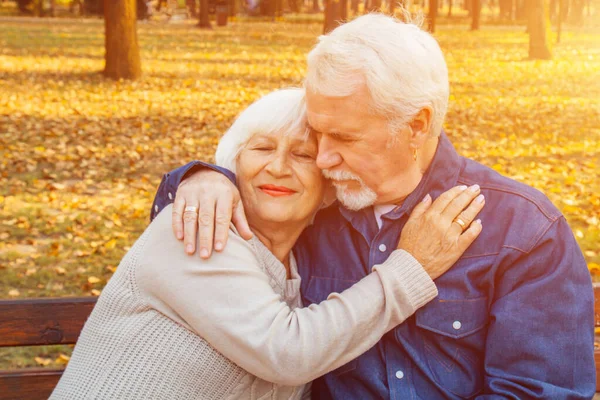 Feliz anciano y mujer sentados en un banco en el día de otoño. Pareja mayor relajada sentada en un banco del parque. Abuelo besa suavemente a la abuela en la frente . — Foto de Stock