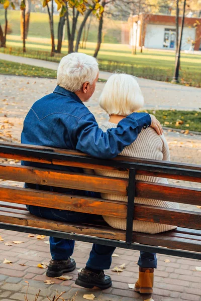 Happy elderly man and woman sitting on a bench in autumn day. Relaxed senior couple sitting on a park bench. Grandfather gently kisses grandmother on the forehead. — Stock Photo, Image